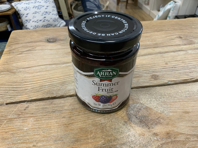 A jar of Summer Fruit Extra Jam sits on a wooden table. The label features strawberries, raspberries, and blackberries, and the lid says "REJECT IF CENTRE BUTTON CAN BE DEPRESSED.