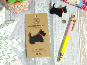 The Scottie Dog Wooden Brooch — All That Glitters, hand-painted and celebrating Scottish heritage, is displayed on a labeled card with a matching brooch, coin, pink-and-yellow pen, and patterned decorative items nearby.