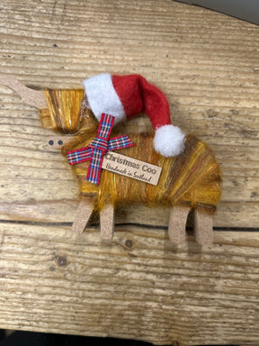 The Hairy Coo Christmas Standing Cow is a small decorative Highland cow made of brown and yellow yarn, wearing a red tartan bow and Santa hat. Labeled "Christmas Coo Handmade in Scotland," it’s perfect festive décor for your home.