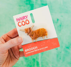 A hand holds a Hairy Coo Brooch, handmade in Scotland and featuring a small Highland cow crafted from brown yarn on a card labeled "BROOCH HANDCRAFTED IN SCOTLAND," set against a green background for Scottish charm.
