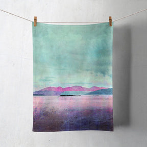 The Arran Over Sound of Bute Tea Towel, made in the UK from Half Panama cotton, hangs from a clothesline with a pastel landscape print of calm water, pink and blue mountains, and a cloudy sky.