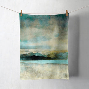 The Skye from Bealach Na Ba Applecross Tea Towel, made from Half Panama cotton, features a blue, green, and beige abstract mountain and water scene. Hung with wooden clothespins on a white wall. Machine washable for convenience.