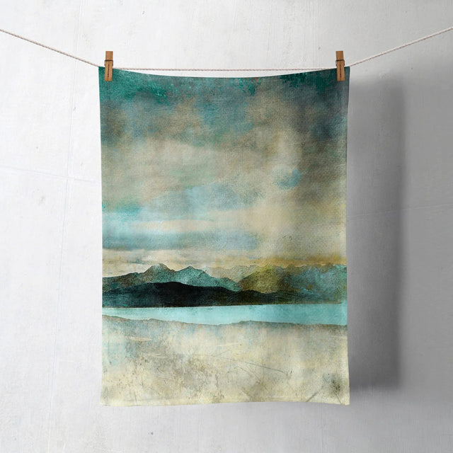 The Skye from Bealach Na Ba Applecross Tea Towel, made from Half Panama cotton, features a blue, green, and beige abstract mountain and water scene. Hung with wooden clothespins on a white wall. Machine washable for convenience.
