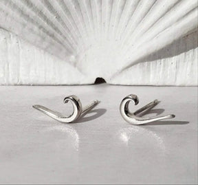 Tidal Wave Earrings – small sterling silver earrings shaped like ocean waves, shown on a white surface with a scallop shell in the background.