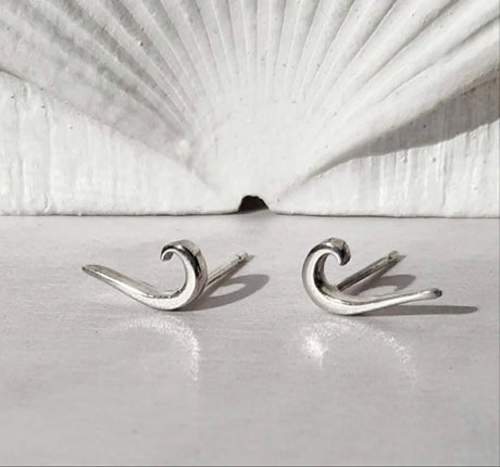 Tidal Wave Earrings – small sterling silver earrings shaped like ocean waves, shown on a white surface with a scallop shell in the background.