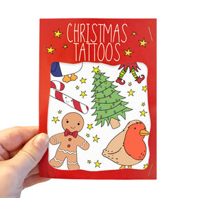 A hand holds a red card labeled "Christmas Transfer Tattoos," displaying festive temporary tattoos such as a gingerbread man, robin, decorated Christmas tree, candy cane, and yellow stars—ideal for spreading holiday cheer.
