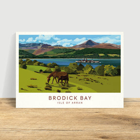 The Catriona Tod Brodick Bay Print Landscape depicts two horses grazing on grassy hills by the water, with a ferry and mountains in the background beneath a blue sky with clouds.