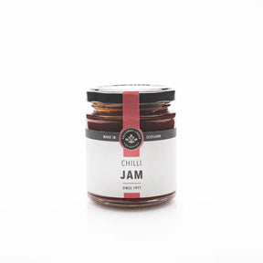 A glass jar of Chilli Jam with a black lid and white label featuring a red stripe, brand logo, and "Chilli Jam." Ideal for adding heat to cheese boards. The jar is shown against a plain white background.