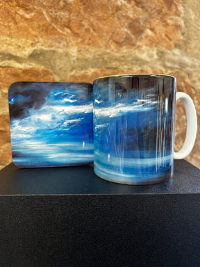 The Blue Yonder Mug and its matching coaster, both with a vibrant blue and white abstract cloud design, are displayed against a textured brown stone background.