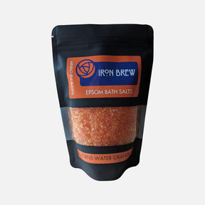 A black and orange resealable pouch labeled "Iron Brew Epsom Salts" by Glasgow Soap Company shows visible orange crystals inside. The text notes "Turns Water Orange." Enjoy a unique soak with these Epsom bath salts.