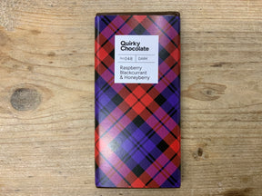 The Raspberry, Blackcurrant and Honeyberry Dark Chocolate Bar has a red and purple tartan-patterned wrapper and features Scottish freeze-dried berries in dark chocolate, presented on a wooden surface.