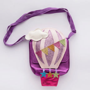 The Children’s Hot Air Balloon Bag is a small purple bag shaped like a hot air balloon, decorated with glitter, vibrant pennant flags, a white cloud, and three stars at the bottom.