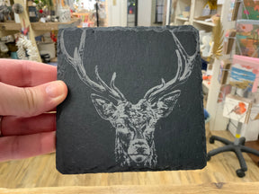 A hand displays the Single Slate Coaster - Stag Prince, a square black slate coaster engraved with an intricate stag head and large antlers, with colorful store shelves visible in the background.