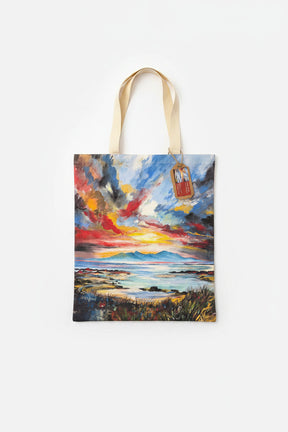 Arran Sunset Tote Bag with beige handles displays a vibrant painting of a dramatic coastal sunset, featuring colorful clouds, water, and grassy foreground. Includes a small tag on one handle.