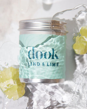 A jar of Dook x Lind & Lime Bath Soak with a light green label sits partially underwater, its rippling reflections evoking citrus bath soak inspiration, while two yellow objects rest nearby.