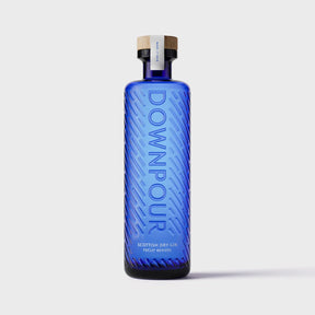 A tall, rectangular blue glass bottle of Downpour Scottish Dry Gin 70cl with a diagonal embossed texture and a wooden stopper, featuring a label that reads “DOWNPOUR” vertically.