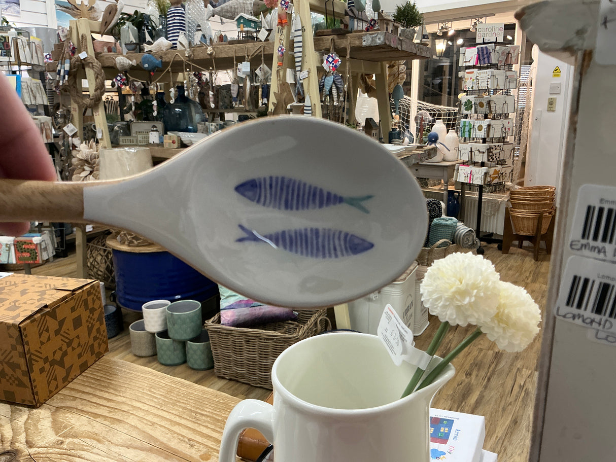 An Enamelled Wooden Spoon with Fish is held above a display table in a gift shop surrounded by assorted home decor items and flowers.