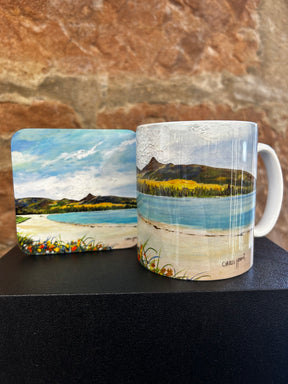 The Goatfell from Brodick Beach Mug and matching coaster, adorned with a painted landscape of mountains, a blue lake, and grassy shores, are displayed against a textured stone background.