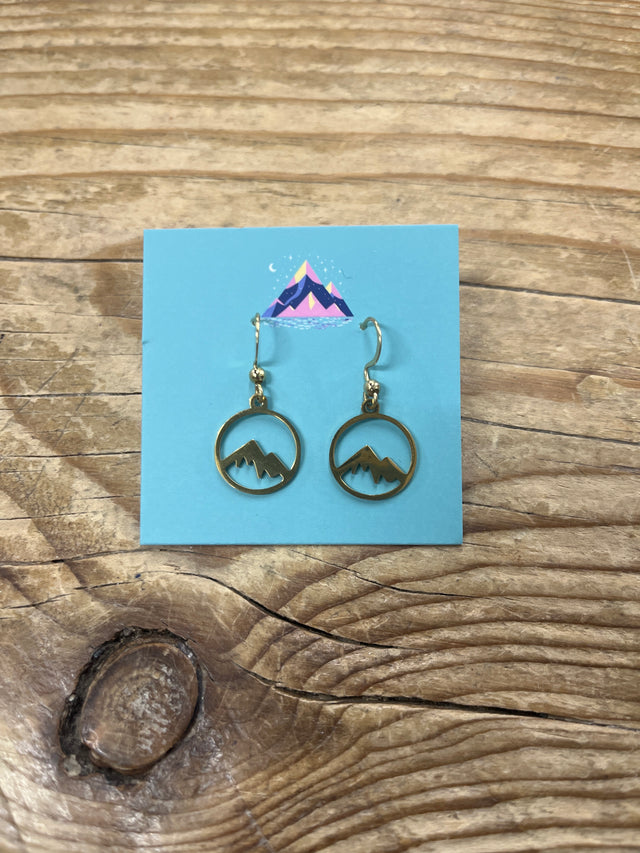 Arran Wishes Arran Earrings Gold, featuring intricate mountain designs within circular gold frames, are showcased on a blue card adorned with a vibrant mountain illustration and set atop a wooden surface. These hypoallergenic earrings are crafted with durable steel hooks for comfortable, everyday wear.