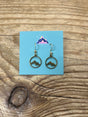 Arran Wishes Arran Earrings Gold, featuring intricate mountain designs within circular gold frames, are showcased on a blue card adorned with a vibrant mountain illustration and set atop a wooden surface. These hypoallergenic earrings are crafted with durable steel hooks for comfortable, everyday wear.