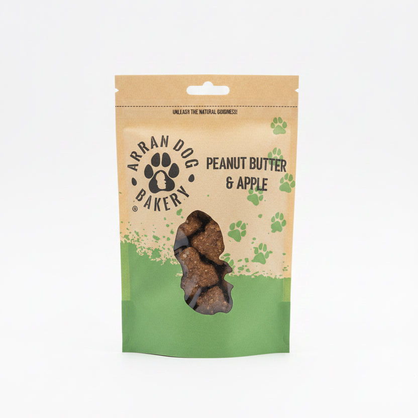 A brown and green pouch of Apple & Peanut Butter Arran Dog Biscuits, featuring a paw print logo and clear window displaying bone-shaped treats with apple inside.
