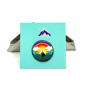 The Arran Wishes Ferry Pin is a round badge featuring a rainbow, ocean waves, and a central ship. Presented on a teal card with a mountain illustration, the unique pin stands upright with its attached metal clip.