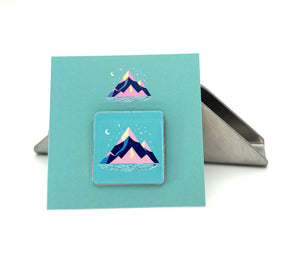 The Arran Wishes Mountain Logo Enamel Pin is a square badge with a stylized pink, purple, and blue mountain scene, displayed on a turquoise card. Ideal for nature pin collectors. A metal stand is visible in the background.