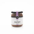 A glass jar of Fig Chutney with a black lid, purple ribbon, and a white label reading "Fig Chutney for Cheese" is the perfect cheese board condiment, shown against a plain white background.