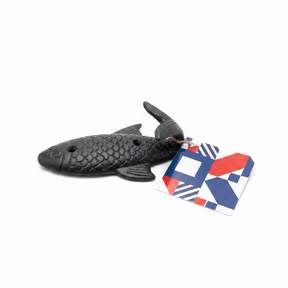 A black Fish wall hook metal with scale details and two holes rests on a white surface, attached to a geometric-patterned tag in blue, red, and white.