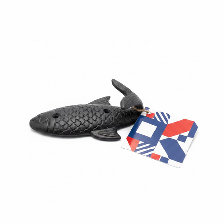 A black Fish wall hook metal with scale details and two holes rests on a white surface, attached to a geometric-patterned tag in blue, red, and white.