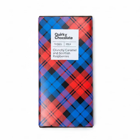 A chocolate bar called "Crunchy Caramel and Scottish Raspberries Chocolate" sits on a wooden surface, wrapped in red, blue, and black tartan. The label reads: "Quirky Chocolate, Crunchy Caramel and Scottish Raspberries, Milk.