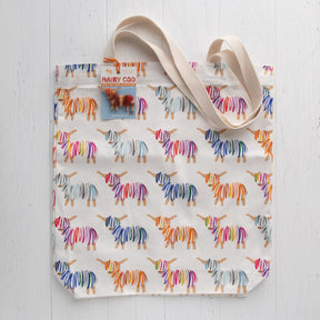 The Hairy Coo Tote Bag is a cream-colored cotton bag made in the UK, featuring playful orange, blue, pink, and gray Hairy Coo Print designs with wide beige handles, shown on a white wooden surface.