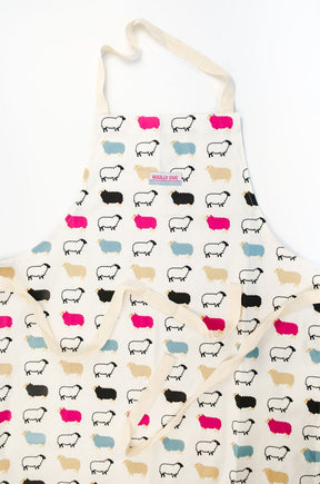 The Woolly Ewe Apron features quirky cartoon sheep in black, beige, pink, and turquoise. This Scottish kitchen apron includes a neck strap, long waist ties, and a "WOOLLY EWES" label near the top center.