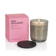 The Max Benjamin Kyoto Blossom Candle, in a gray glass jar with a gold logo, burns beside its pink box. A black lid sits in front, all set against a white background.