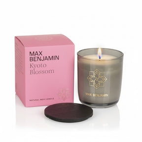 The Max Benjamin Kyoto Blossom Candle, in a gray glass jar with a gold logo, burns beside its pink box. A black lid sits in front, all set against a white background.