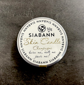 A Siabann Champagne Skin Candle 50g rests on a dark surface, labeled "burn me, melt me, pour me!" in gold and black. This round candle melts into a nourishing massage oil made from natural oils.