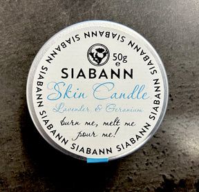 A round tin labeled "Siabann Lavender and Geranium Skin Candle 50 g" with "burn me, melt me, pour me!" on the lid rests on a dark surface. The candle features natural oils and the brand name circles the edge in black text.