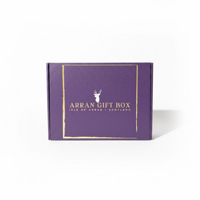 A box with a purple shade and golden text reading "Arran Gift Box - Medium Size Gift Box, Isle of Arran, Scotland" rests on rocky terrain near the water's edge, promising a delightful experience of Scottish delicacies. The backdrop features a serene lake, distant mountains, and a bright blue sky.