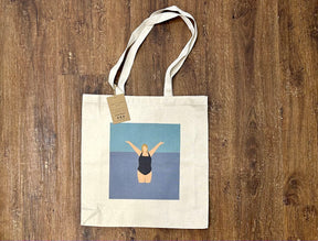 The "I Love the Water Tote Bag" by Fiddy and Mabel is a beige eco-friendly cotton tote with long handles, thick canvas, and a brown tag. It shows a person in a black swimsuit in water with raised arms against a blue background.