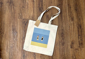 The Chats in the Sea Tote Bag by Fiddy and Mabel, made of thick beige canvas, rests on a wooden floor. It features an illustration of two women in swimsuits on a beach with blue sky and yellow sand, plus a brown tag on one handle.