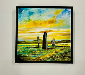 The Standing Stones Ceramic Tile features three standing stones in a grassy field under a dramatic sky, inspired by Arran landscapes, with yellow and blue clouds and distant hills on the horizon.