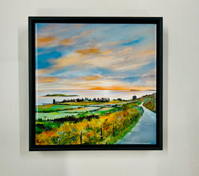 The Road to Kildonan Ceramic Tile, inspired by Charles Herbert, depicts a winding road through grassy fields and distant trees beneath a dramatic sunset sky, and is displayed on a plain white wall.