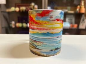 The Arran Sunset Mug is a ceramic mug adorned with a colorful painted landscape of mountains, a lake, and a vibrant sunset sky. It is displayed on a white surface with blurred shelves in the background.