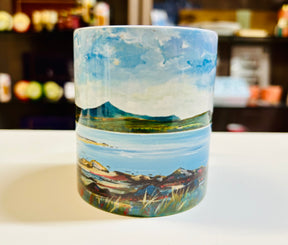 The Brodick Goatfell Mug is a ceramic mug adorned with a vibrant landscape painting of a lake, mountains, grassy shore, and blue sky. It rests on a white surface with blurred shelves in the background.