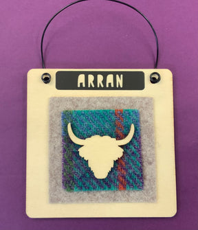 The Arran Wooden Hanging Sign — All That Glitters is handmade in Scotland with a wire handle, "ARRAN" label, a highland cow head cutout on multicolored textured fabric, set against a bold purple background.