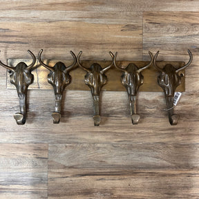 The Brass Buffalo Hooks is a wall-mounted rack featuring five metal hooks shaped like steer skulls, displayed against a wooden floor.