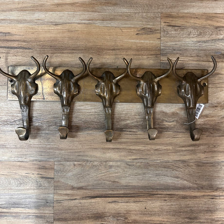The Brass Buffalo Hooks is a wall-mounted rack featuring five metal hooks shaped like steer skulls, displayed against a wooden floor.