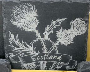 A Single Slate Coaster featuring an engraving of two Scottish thistle flowers and leaves, with a banner across the stems that reads “Scotland.”.