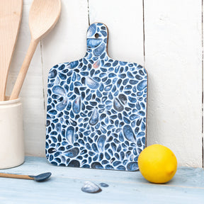 The Mussel Chopping Board features a blue pebble and mussel shell design and stands upright on a blue wooden surface beside a ceramic jar of wooden utensils and a lemon, adding coastal charm. White wooden planks complete the backdrop.