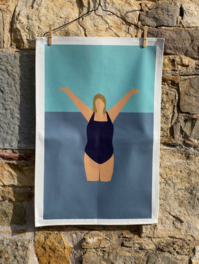 The "I Love the Water Tea Towel" features a minimalist illustration of a woman in a dark swimsuit, arms raised in water—a tribute to wild swimming. The design hangs on a line against stone, with blue and teal cotton-inspired background details.